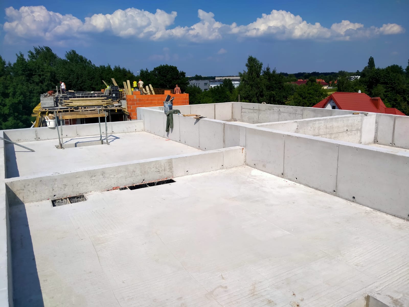 Świeżo wylana płyta fundamentowa z betonowymi ścianami działowymi, widoczne szalunki i pracownicy w tle, słoneczny dzień.