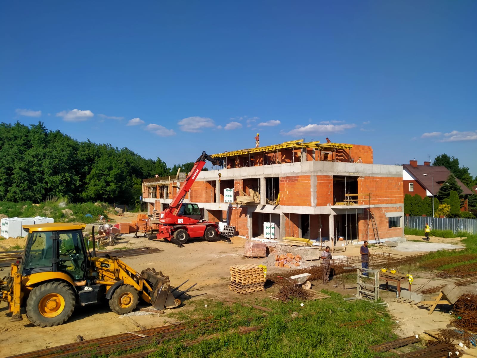 Wznoszenie dwukondygnacyjnego budynku z czerwonej cegły, widoczny żuraw teleskopowy i ładowarka kołowa, szalunki na stropie, pracownicy na budowie, w tle dom jednorodzinny i drzewa.