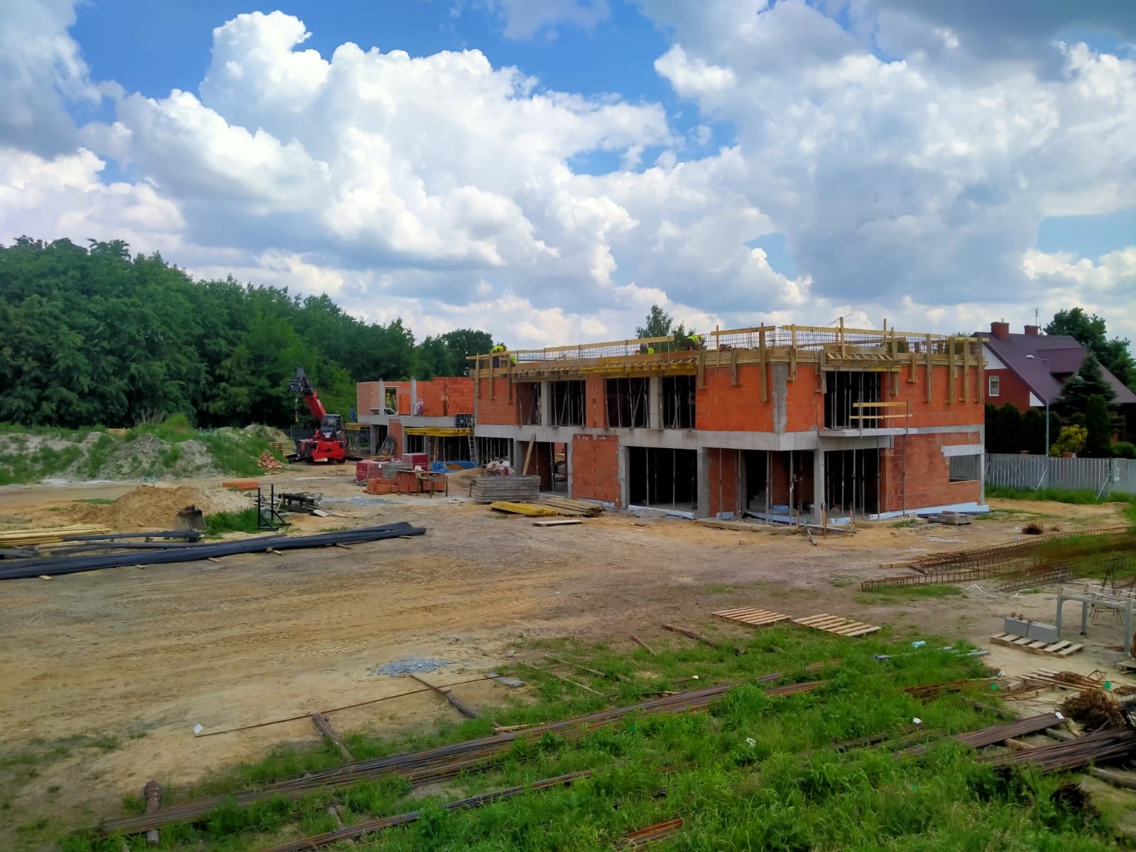 Wznoszenie dwukondygnacyjnego budynku żelbetowego z czerwonej cegły, widoczny dźwig na placu budowy, stal zbrojeniowa i drewniane szalunki na górze, w tle drzewa i błękitne niebo z białymi chmurami.