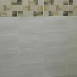 Wykończona ściana łazienki z jasnymi, prostokątnymi płytkami ceramicznymi ułożonymi poziomo, z dekoracyjnym pasem mozaiki kwadratowej w odcieniach beżu i brązu u góry.