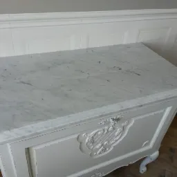 Biały marmurowy blat na komodzie w stylu vintage z dekoracyjnymi ornamentami, widoczne ślady użytkowania i delikatna patyna.