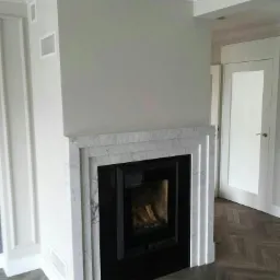 Nowoczesny kominek z obudową z białego marmuru z czarnym wkładem, widoczny parkiet w jodełkę i białe drzwi w tle.