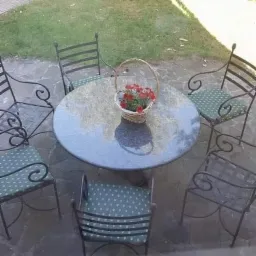 Okrągły stół z ciemnego kamienia na tarasie otoczony metalowymi krzesłami z zielonymi poduszkami w kropki, koszyk z czerwonymi kwiatami na stole, widok zza szyby.