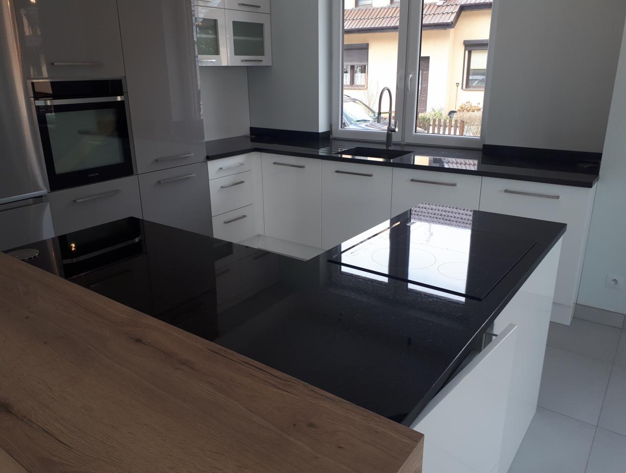 Nowoczesna kuchnia z białymi szafkami i czarnym blatem, wyspa kuchenna z płytą indukcyjną, drewniany blat na wyspie, widok na dom z okna.