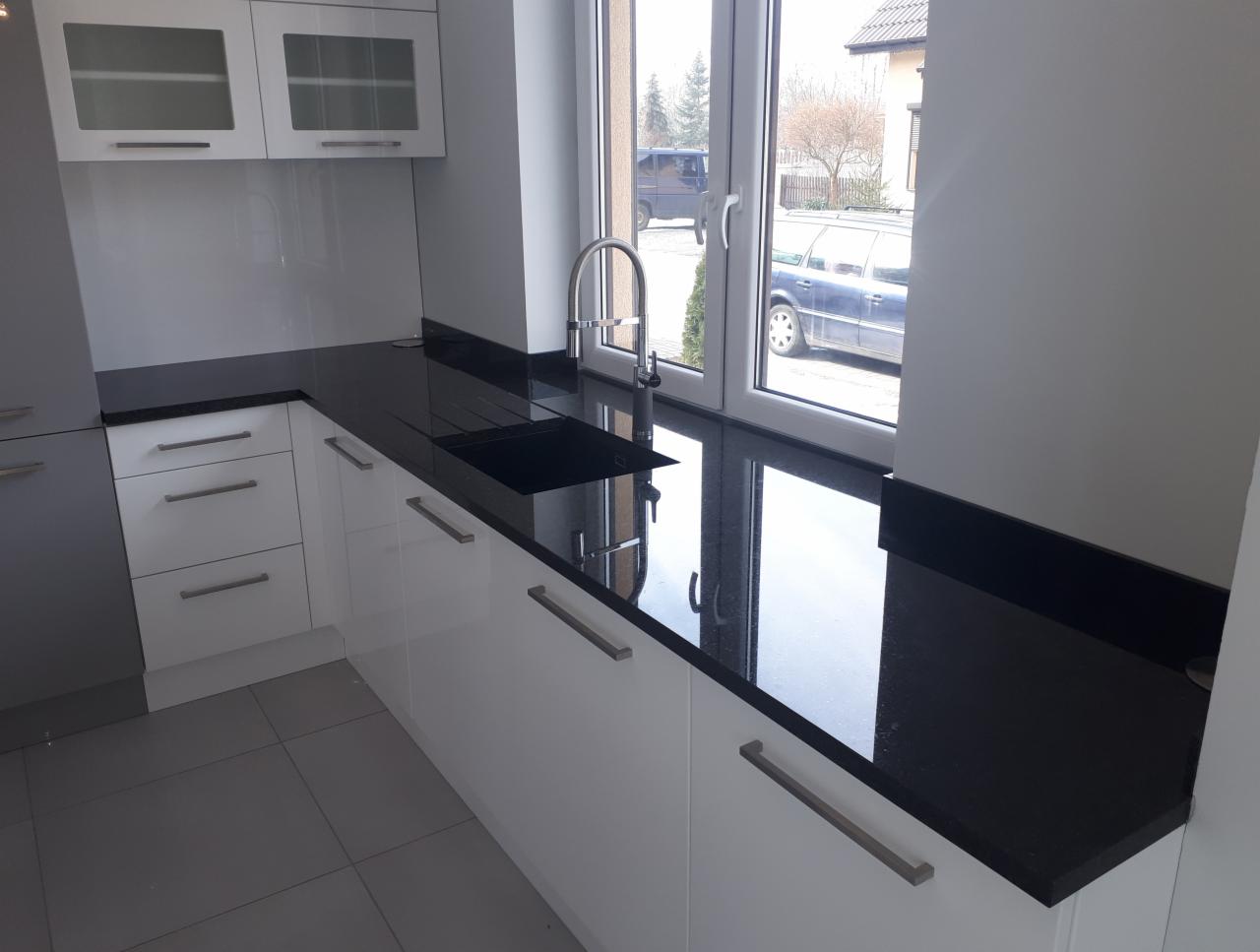 Czarny, błyszczący blat kuchenny z wbudowanym zlewem, zamontowany w nowoczesnej kuchni z białymi szafkami i szarą podłogą. Okno z widokiem na ulicę i zaparkowane samochody w tle.