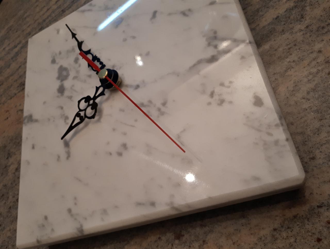 Kwadratowy zegar z białego marmuru z szarymi żyłkami, wskazówki czarne z czerwoną sekundą, leżący na blacie.