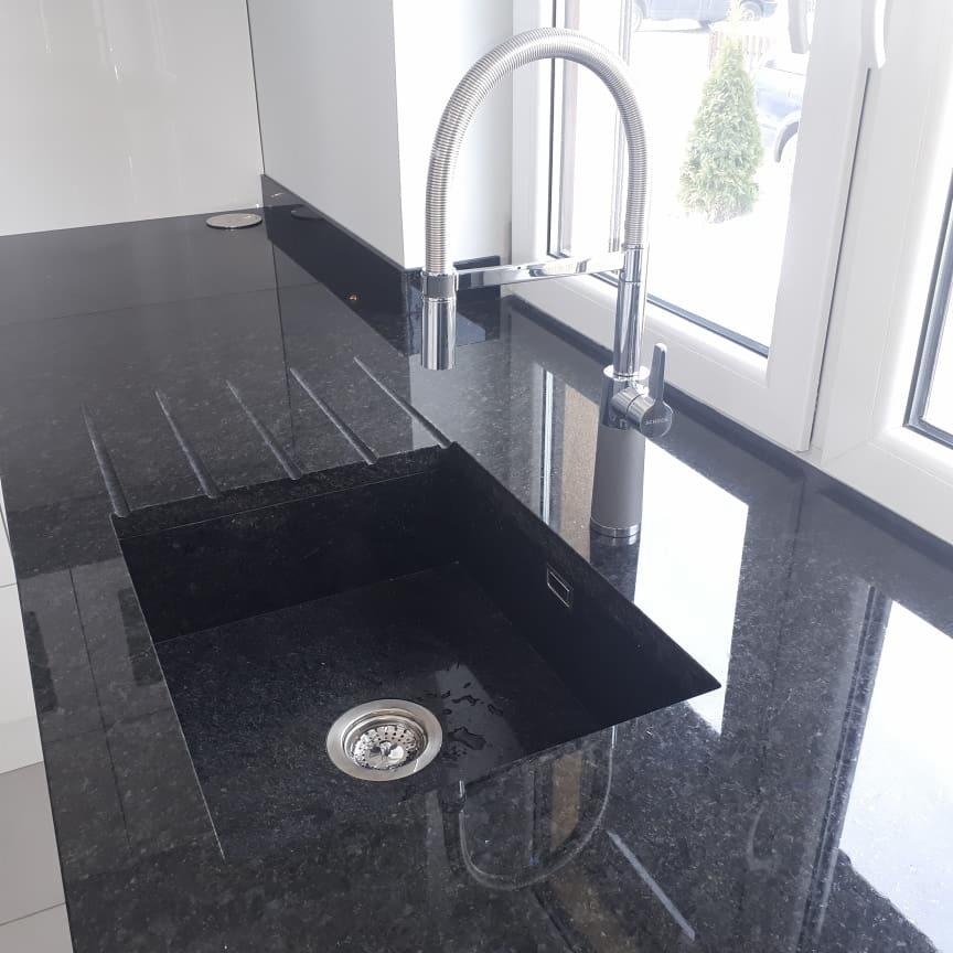 Czarny, polerowany blat kuchenny z granitową wbudowaną umywalką i chromowaną baterią z elastyczną wylewką, widok z góry, jasne wnętrze kuchni.