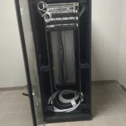 Otwarta szafa rack z zamontowanymi urządzeniami sieciowymi, widoczne kable i patch panel, w tle ściana z białych bloczków.