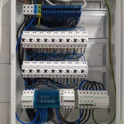 Otwarta skrzynka rozdzielcza z licznymi bezpiecznikami, przewodami i listwami zaciskowymi, zamontowana na ścianie z bloczków betonowych.