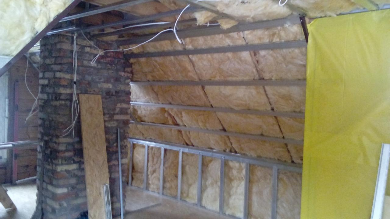 Docieplenie poddasza wełną mineralną, widoczna konstrukcja metalowa, fragment komina z cegły i wystające przewody elektryczne.