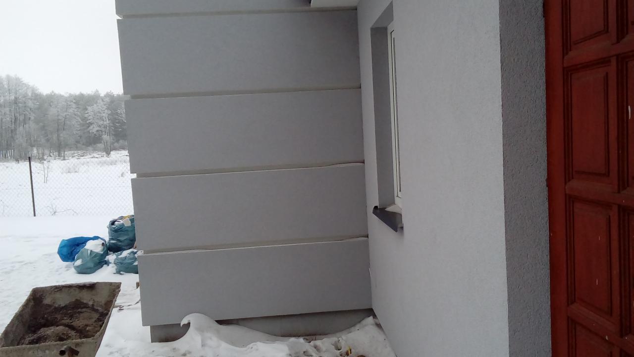 Docieplenie elewacji budynku z widocznymi panelami izolacyjnymi, oknem i fragmentem drewnianych drzwi wejściowych w zimowej scenerii.