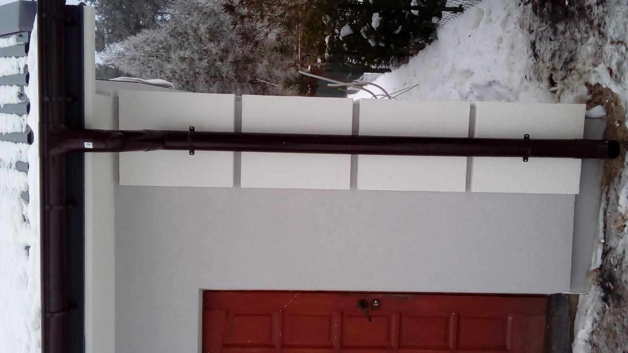 Docieplenie ściany budynku z widocznymi panelami izolacyjnymi, brązową rynną i częściowo ośnieżonym otoczeniem, z czerwoną, drewnianą stolarką drzwiową.