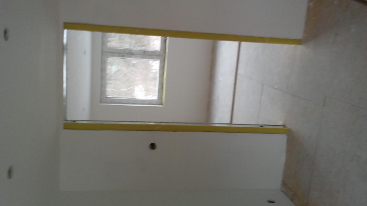 Fragment ściany z otworem okiennym, zabezpieczonym żółtą taśmą malarską, przygotowywanej do wykończenia, widoczne ślady po szpachlowaniu.