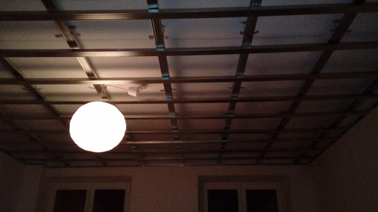 Szkielet sufitu podwieszanego z widoczną metalową konstrukcją kratową i częściowo zamontowanymi płytami, oświetlony kulistą lampą wiszącą, widoczne okna w tle.