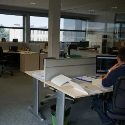 Pracownik biurowy siedzi przy biurku przed komputerem, analizując dane; widoczne segregatory i dokumenty na blacie w jasnym open space.