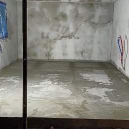 Rafał Śmist Usługi Remontowo-Budowlana - Surowe wnętrze w trakcie prac wykończeniowych: świeżo wylana, mokra posadzka, przygotowane ściany z widocznymi śladami napraw, zabezpieczone okna folią, widoczne instalacje elektryczne...