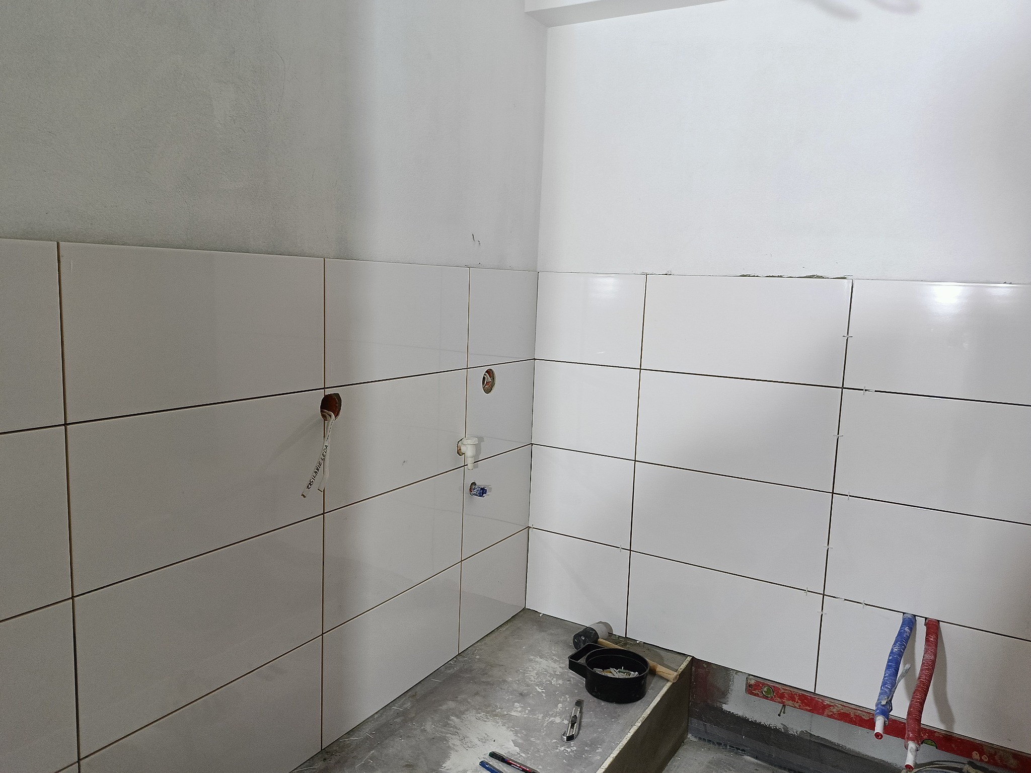 Wnętrze łazienki w trakcie kładzenia białych, prostokątnych płytek na ścianach; widoczne wystające rury, poziomica, pojemnik z narzędziami i niedokończona podłoga.