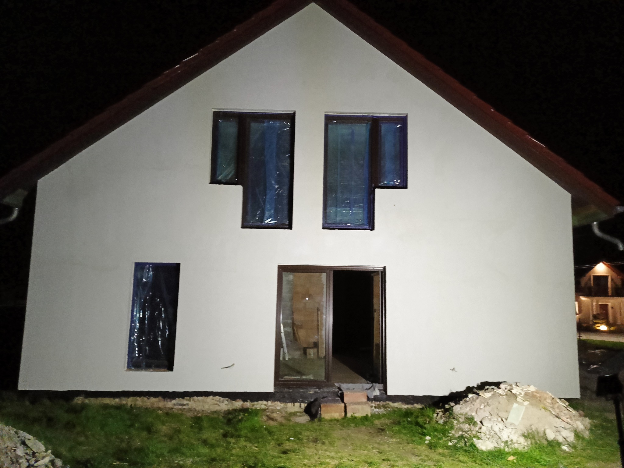 Nocne ujęcie elewacji domu jednorodzinnego w trakcie prac wykończeniowych, okna zabezpieczone folią, widoczne ślady gruzu przy wejściu.