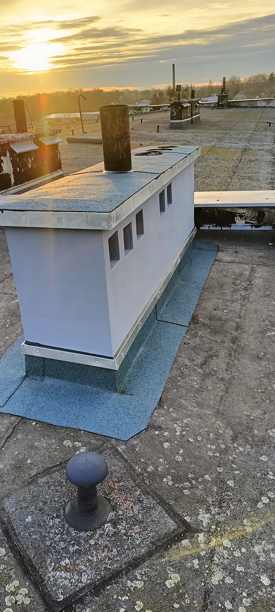 Szary, odnowiony komin wentylacyjny z małymi otworami na płaskim dachu pokrytym papą w blasku wschodzącego słońca.