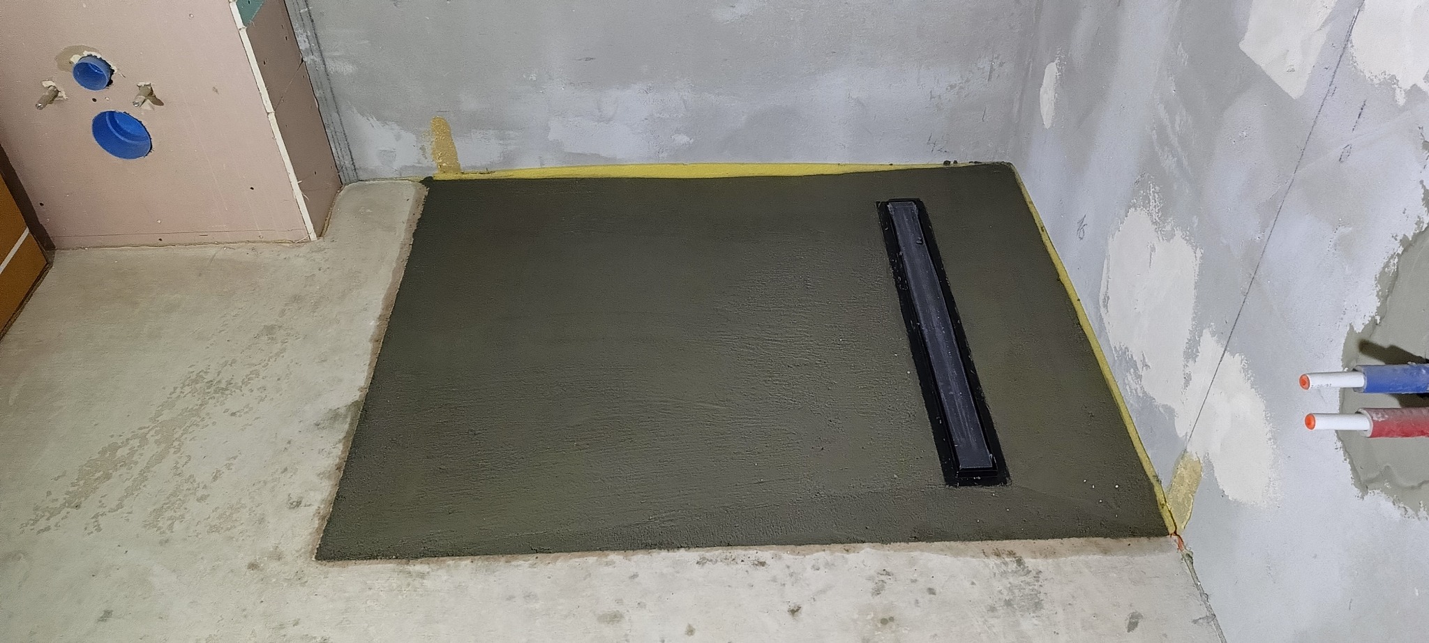 Wylewka pod prysznic z odpływem liniowym, świeżo wykonana, zabezpieczona żółtą taśmą, widoczne rury instalacji wodnej w ścianie, surowy stan ścian.