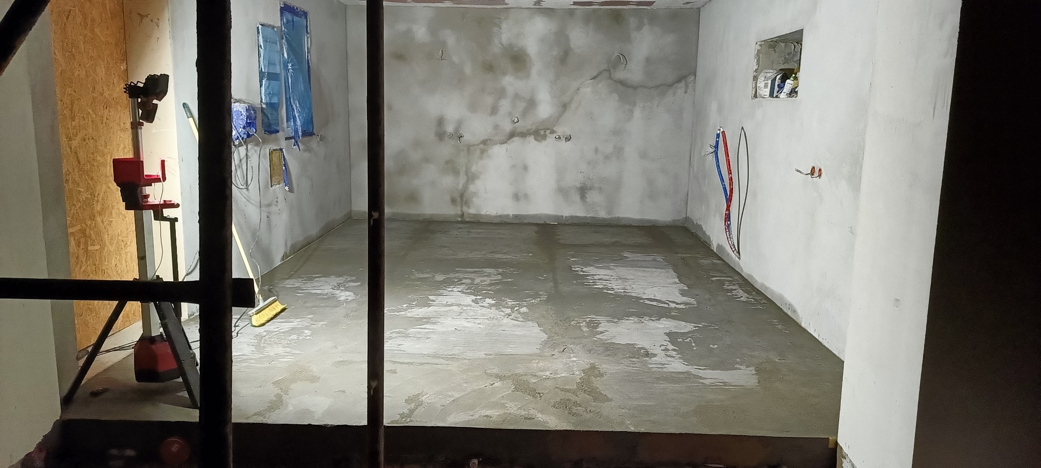 Surowe wnętrze w trakcie prac wykończeniowych: świeżo wylana, mokra posadzka, przygotowane ściany z widocznymi śladami napraw, zabezpieczone okna folią, widoczne instalacje elektryczne...