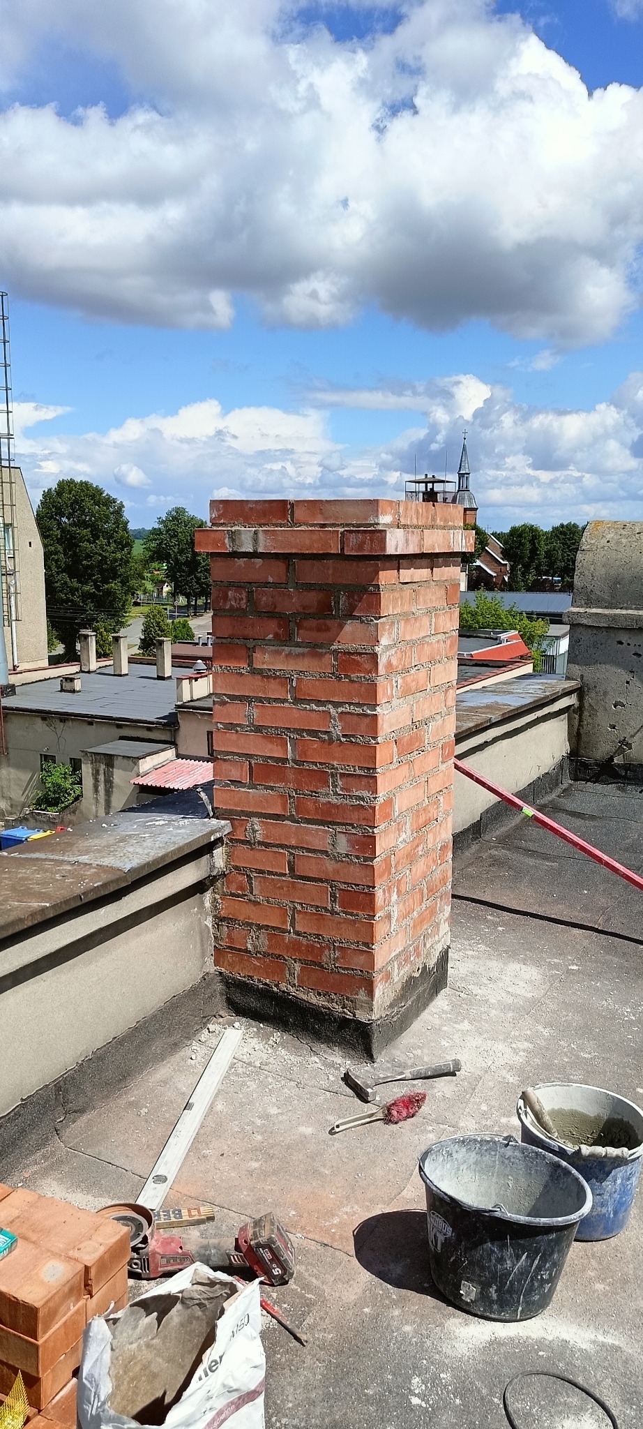 Murowany komin z czerwonej cegły na dachu, widoczne narzędzia murarskie, wiaderka z zaprawą, poziomica, w tle zabudowa miejska i błękitne niebo z chmurami.