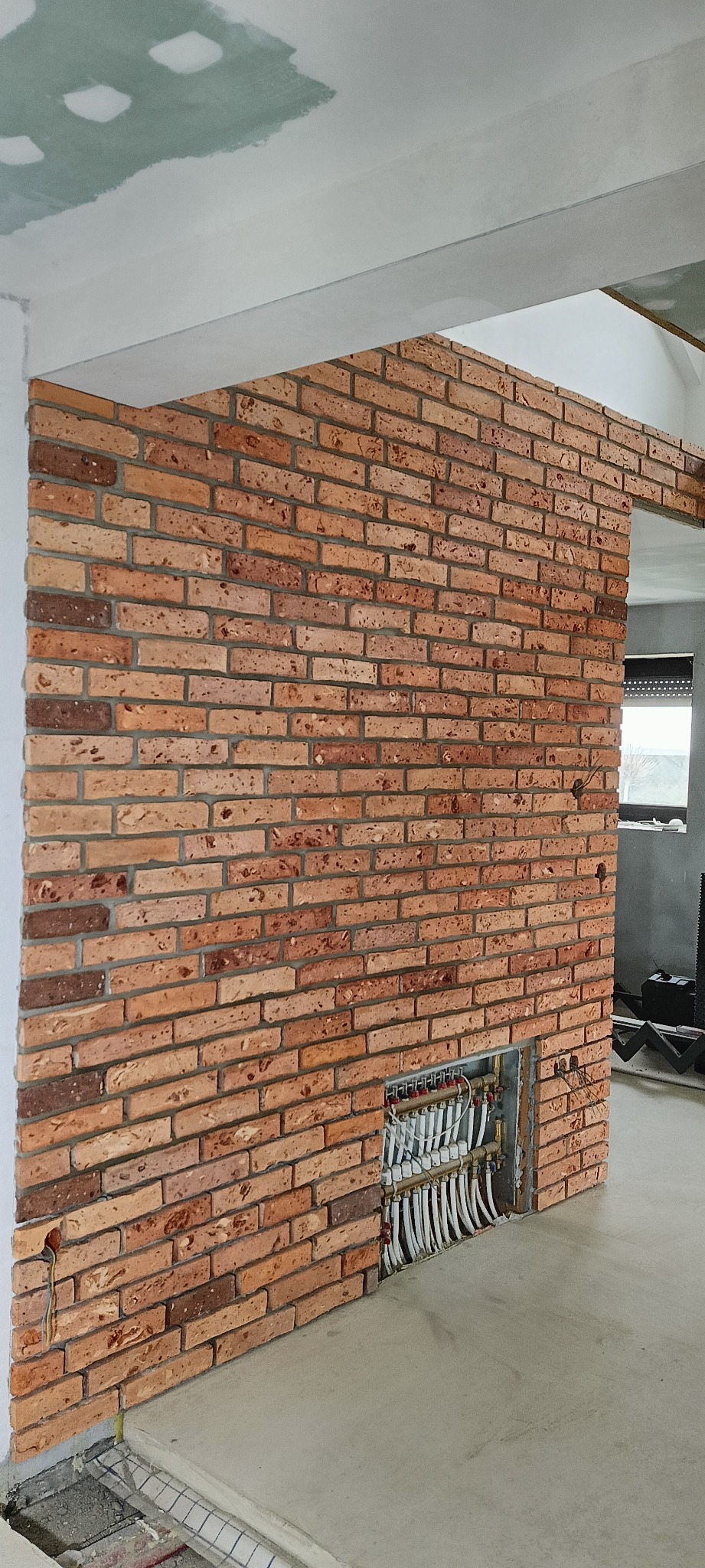 Ściana z czerwonej cegły klinkierowej w surowym wnętrzu, widoczny fragment instalacji ogrzewania podłogowego w dolnej części ściany, sufit w trakcie wykańczania.