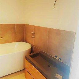 PABLO Paweł Wasąg - Nowoczesna łazienka w trakcie wykańczania: wolnostojąca wanna z baterią, drewniana szafka z szarą umywalką i brązowe płytki na ścianie, widoczne wystające przewody elektryczne.