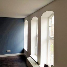 PABLO Paweł Wasąg - Pokój po odświeżeniu z ciemnoniebieską ścianą, trzema niszami okiennymi z białymi ościeżami i ciemną podłogą z listwami przypodłogowymi.