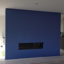 PABLO Paweł Wasąg - Świeżo pomalowana, intensywnie niebieska ściana z wbudowanym, poziomym biokominkiem w nowoczesnym wnętrzu, widoczne zabezpieczenia podłogi i fragmenty innych ścian w trakcie prac.