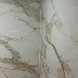 Ściana wyłożona białymi płytkami imitującymi marmur z delikatnymi brązowymi żyłami, widoczny narożnik i fragment baterii.