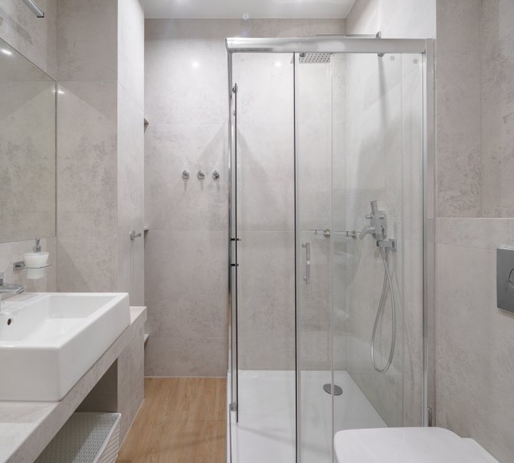 Nowoczesna łazienka z kabiną prysznicową ze szklanymi drzwiami przesuwnymi, umywalką nablatową o kwadratowym kształcie i jasnymi płytkami imitującymi kamień na ścianach.