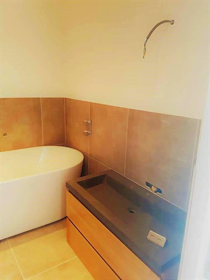 Nowoczesna łazienka w trakcie wykańczania: wolnostojąca wanna z baterią, drewniana szafka z szarą umywalką i brązowe płytki na ścianie, widoczne wystające przewody elektryczne.