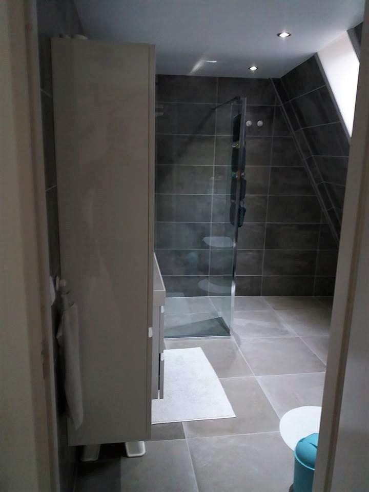 Nowoczesna łazienka z kabiną prysznicową ze szklanymi drzwiami, szarą glazurą, szafką z umywalką i ręcznikiem na wieszaku, oświetleniem punktowym i skośnym sufitem.