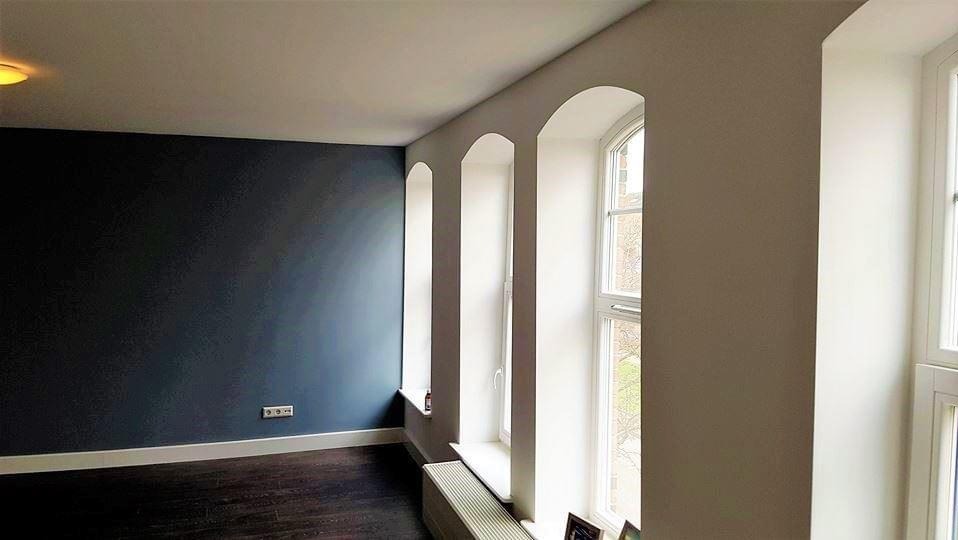 Pokój po odświeżeniu z ciemnoniebieską ścianą, trzema niszami okiennymi z białymi ościeżami i ciemną podłogą z listwami przypodłogowymi.