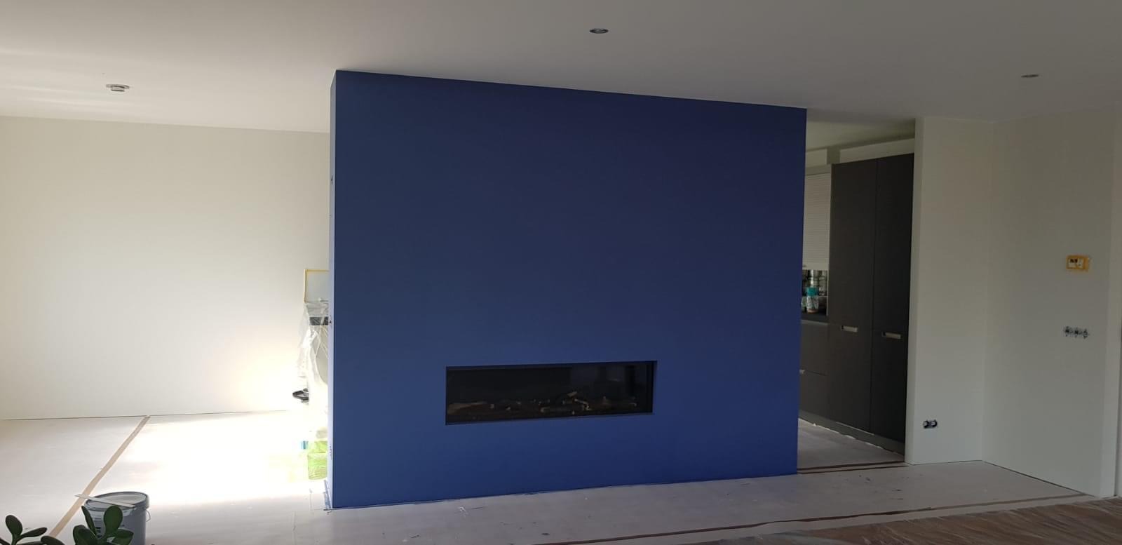 Świeżo pomalowana, intensywnie niebieska ściana z wbudowanym, poziomym biokominkiem w nowoczesnym wnętrzu, widoczne zabezpieczenia podłogi i fragmenty innych ścian w trakcie prac.