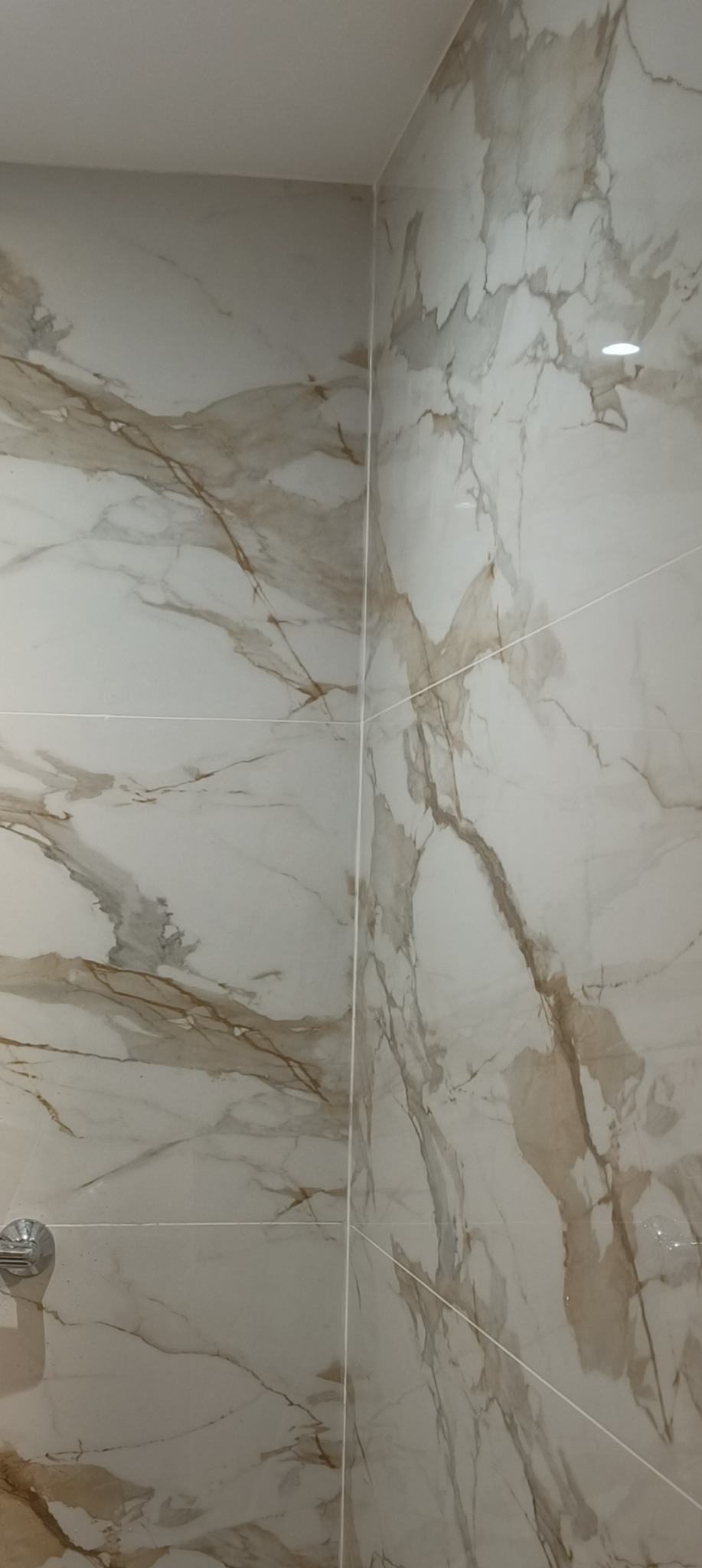 Ściana wyłożona białymi płytkami imitującymi marmur z delikatnymi brązowymi żyłami, widoczny narożnik i fragment baterii.