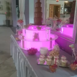 Stół z fontanną czekoladową, podświetlany na różowo, udekorowany babeczkami, owocami i popcornem, w tle lustro odbijające salę bankietową.