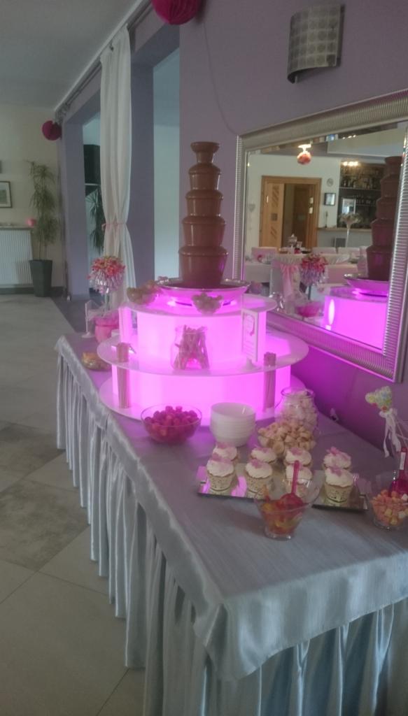 Stół z fontanną czekoladową, podświetlany na różowo, udekorowany babeczkami, owocami i popcornem, w tle lustro odbijające salę bankietową.