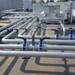 Równoległe rury instalacyjne z zaworami na dachu budynku, widoczne urządzenia klimatyzacyjne w tle.