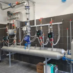Rozbudowana instalacja grzewcza z rurami, zaworami i zbiornikami, widoczne manometry i automatyka, tło stanowią ciemne płytki ceramiczne.