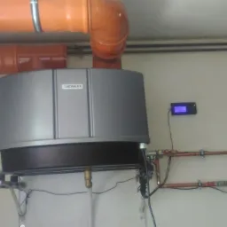 Szary piec gazowy Hewalex z podłączonymi miedzianymi rurami i wyświetlaczem na ścianie, widoczne pomarańczowe rury wentylacyjne.