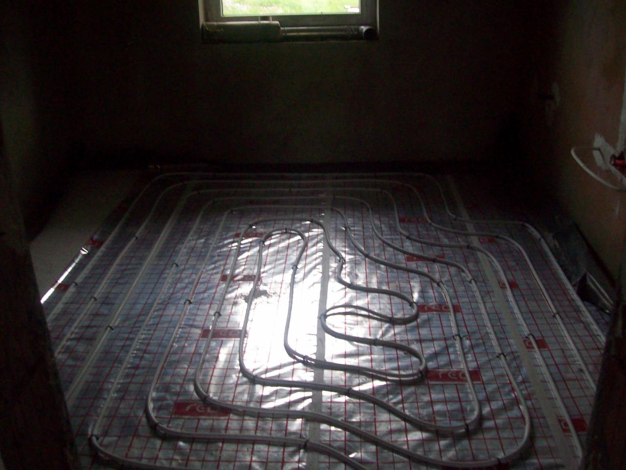 Układanie rur ogrzewania podłogowego na folii z rastrem w słabo oświetlonym pomieszczeniu, widok z góry na instalację przed zalaniem wylewką.