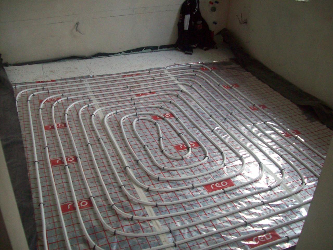 Białe rury ogrzewania podłogowego rozłożone spiralnie na srebrnej folii z czerwoną kratką w trakcie montażu.