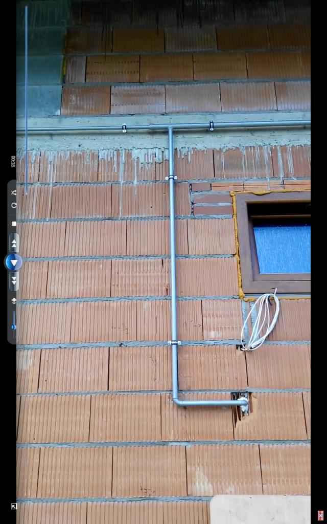 Szara rura instalacyjna przymocowana do ściany z czerwonej cegły, pionowy odcinek rury biegnie w dół, a poziomy odcinek w prawo, obok okna z brązową ramą uszczelnionego żółtą pianką.