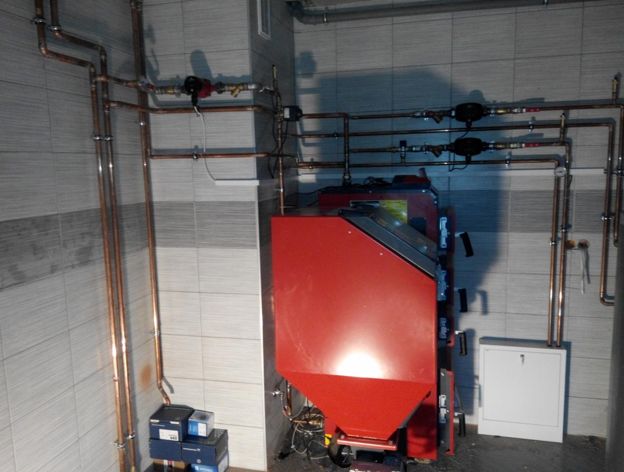 Czerwony piec CO z podajnikiem na ekogroszek, widoczne miedziane rury instalacji grzewczej na tle szaro-białych płytek ceramicznych.