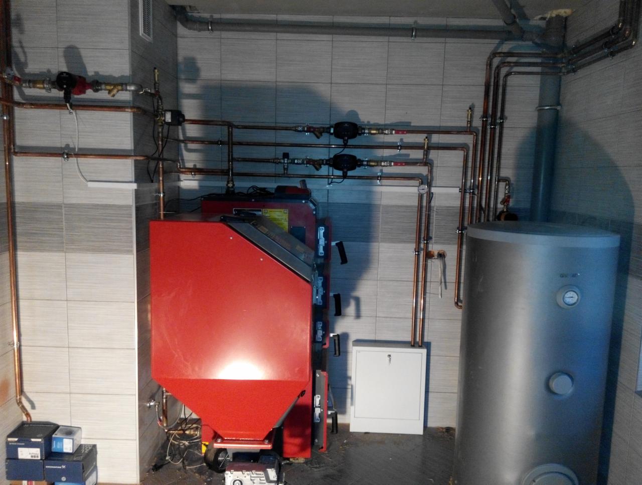 Czerwony piec na paliwo stałe z podajnikiem ślimakowym i srebrny zasobnik ciepłej wody, połączone rurami miedzianymi w kotłowni wyłożonej szarymi płytkami.