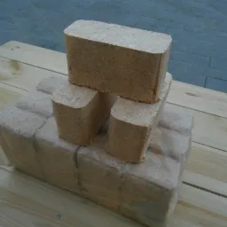 Ułożone na drewnianym blacie kostki sprasowanych trocin w przezroczystym opakowaniu foliowym, trzy kostki ułożone w piramidkę na wierzchu.