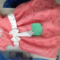 Różowa, koronkowa sukienka dziewczęca z białą satynową kokardą, leżąca na stole z zieloną kartką z zapisanym rozmiarem.