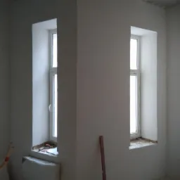 Wnętrze pokoju po nałożeniu jasnoszarego tynku maszynowego, widoczne dwa okna z białymi ramami, grzejnik pod jednym z nich i pionowa listwa przypodłogowa przy ścianie.